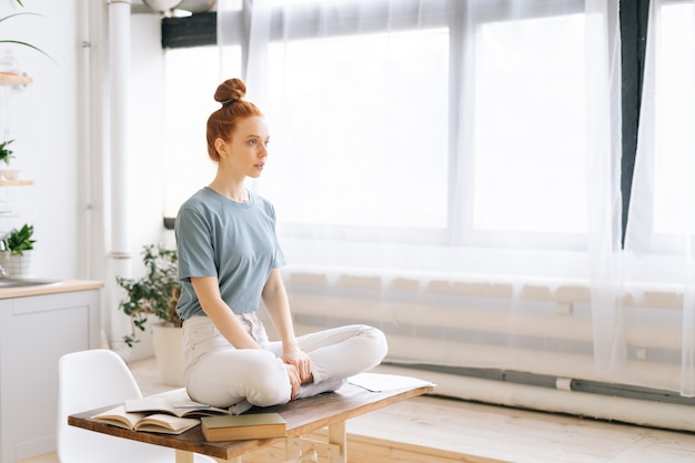 Retrato de uma jovem ruiva sorridente meditando enquanto está sentado na mesa do escritório em casa