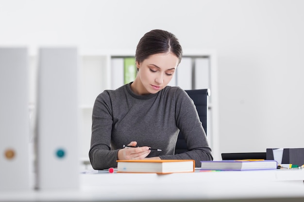 Retrato de uma jovem secretária vestindo um suéter cinza e escrevendo em seu caderno em seu local de trabalho em um escritório branco.