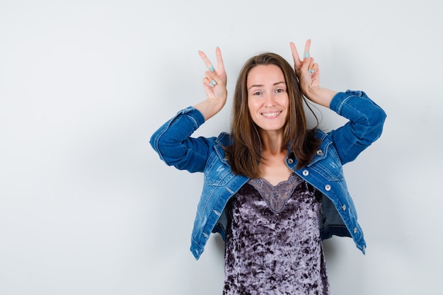 Retrato de uma jovem senhora mostrando o gesto de orelhas de coelho no vestido, jaqueta jeans e olhando a vista frontal engraçada