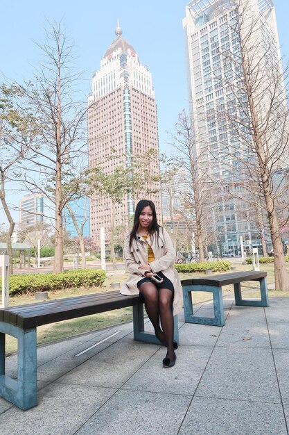 Foto retrato de uma jovem sentada em um banco contra edifícios no parque