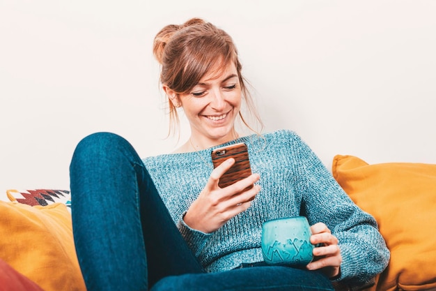 Retrato de uma jovem sentada em um sofá, assistindo no celular e sorrindo - conceito chill out - conceito de comunicação sem fio