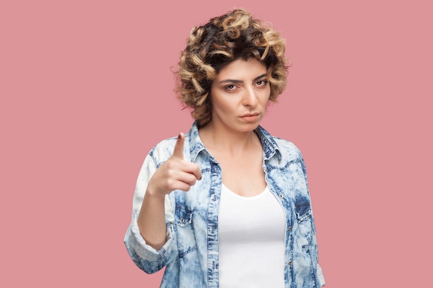 Retrato de uma jovem séria com penteado encaracolado em pé de camisa azul casual, olhando para a câmera com a mão de advertência. tiro de estúdio interno, isolado no fundo rosa.