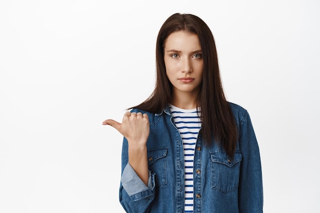 Retrato de uma jovem séria não gosta de smth, apontando para a esquerda e olhando com rosto preocupado e cético, expressa sua descrença ou julgamento, de pé sobre fundo branco.