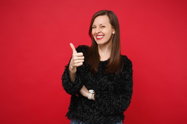 Retrato de uma jovem sorridente alegre em pé de suéter de pele preta, aparecendo o polegar isolado no fundo da parede vermelha brilhante no estúdio. Emoções sinceras de pessoas, conceito de estilo de vida. Simule o espaço da cópia.