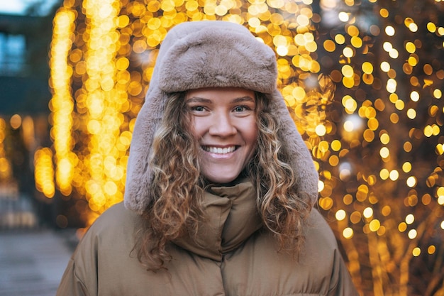 Retrato de uma jovem sorridente com cabelo encaracolado com chapéu de pele na rua de inverno decorada com luzes