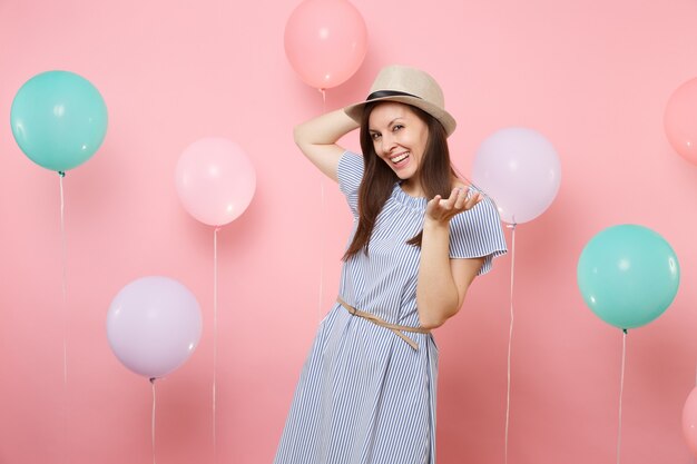 Retrato de uma jovem sorridente com chapéu de palha de verão e vestido azul em pé sobre fundo rosa pastel com balões de ar coloridos. Conceito de emoções sinceras de pessoas de festa de férias de aniversário.