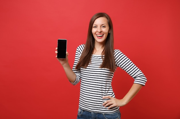 Retrato de uma jovem sorridente com roupas listradas, segurando o telefone móvel com tela vazia preta em branco, isolada em fundo vermelho brilhante. Emoções sinceras de pessoas, conceito de estilo de vida. Simule o espaço da cópia.