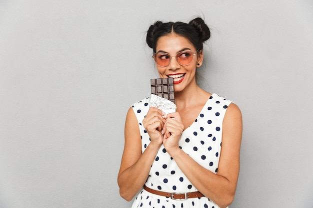 Retrato de uma jovem sorridente com vestido de verão e óculos escuros isolados, segurando a barra de chocolate