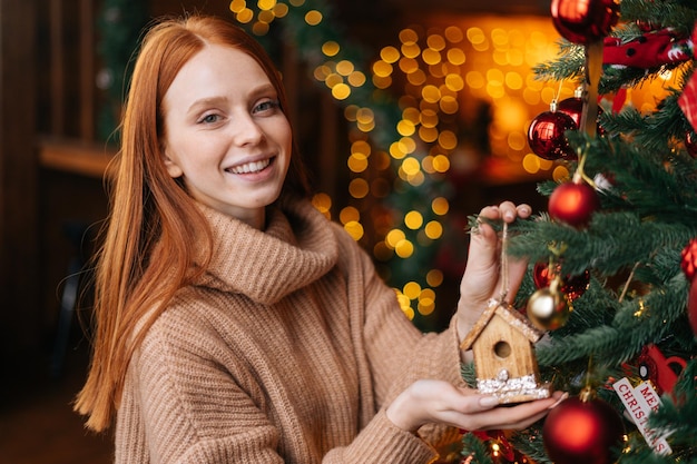 Retrato de uma jovem sorridente decorando um lindo brinquedo de árvore de Natal na aconchegante sala de estar