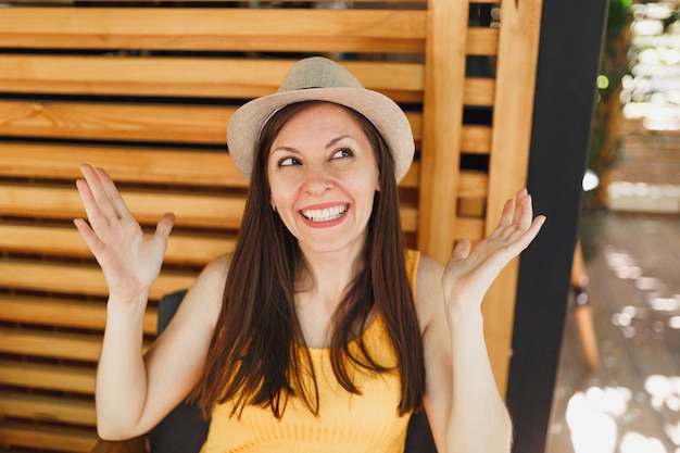 Retrato de uma jovem sorridente e animada com chapéu de palha de verão, camisa amarela, espalhando as mãos na parede de madeira em rua ao ar livre, café, café, verão