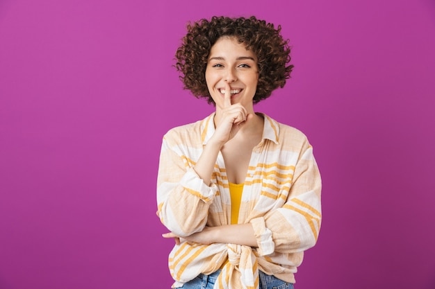 Retrato de uma jovem sorridente e atraente com cabelo castanho encaracolado isolado na parede violeta, mostrando um gesto de silêncio