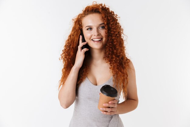 Retrato de uma jovem sorridente e atraente, com cabelo ruivo longo cacheado, isolado, falando no celular enquanto bebe café para viagem