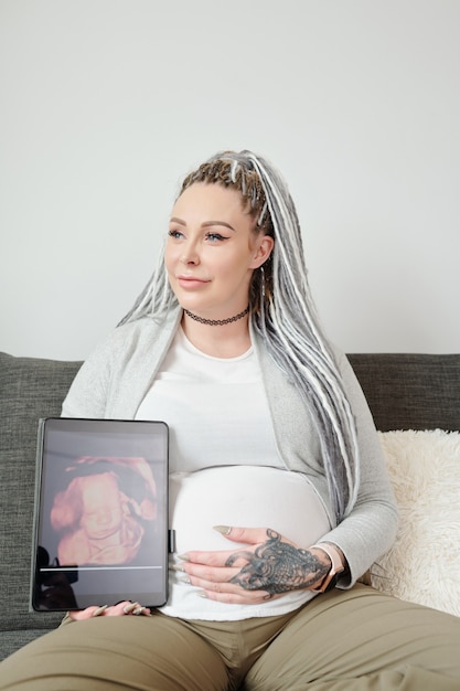 Foto retrato de uma jovem sorridente e feliz com dreadlocks, sentado no sofá e mostrando um retrato de ultrassom de bebê