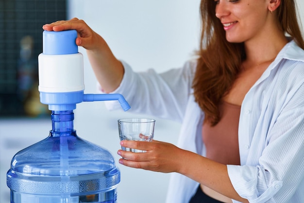 Retrato de uma jovem sorridente e feliz derramando água fresca limpa em vidro de garrafa grande em casa