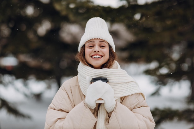 Retrato de uma jovem sorridente e feliz, vestido com cachecol, chapéu e luvas, aproveita o clima de inverno no parque de inverno nevado