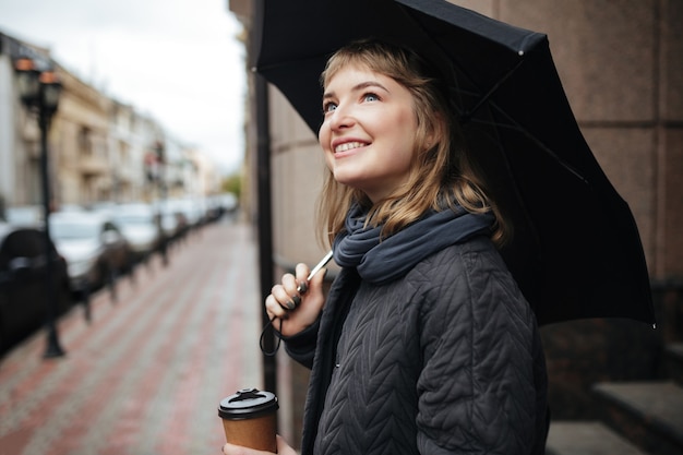 Retrato de uma jovem sorridente em pé na rua com um guarda-chuva preto e café nas mãos e olhando feliz para o lado