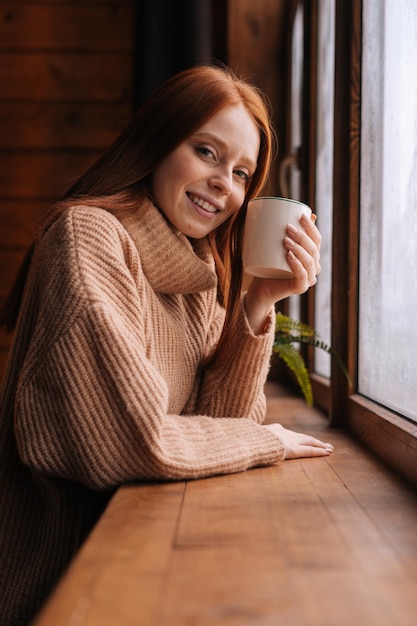 Retrato de uma jovem sorridente em pé perto da janela com uma xícara de café quente e olhando para a câmera