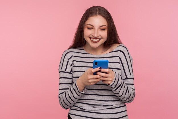 Retrato de uma jovem sorridente feliz digitando no celular, lendo um blog engraçado, conversando na rede social, usando o aplicativo móvel para comunicação on-line. tiro de estúdio interior isolado no fundo rosa