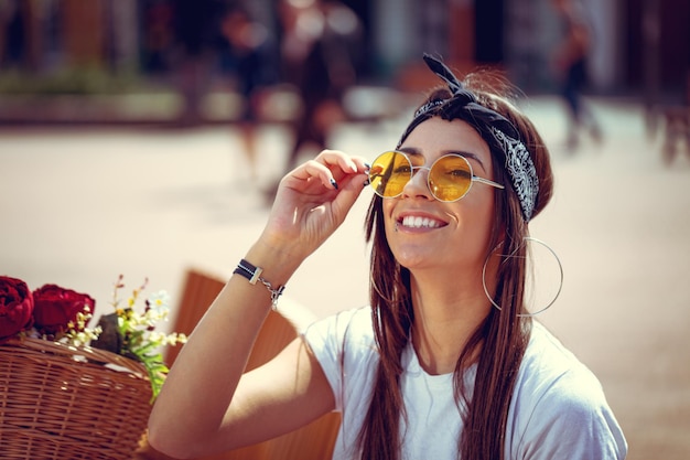 Retrato de uma jovem sorridente feliz que está desfrutando em um dia ensolarado de verão, sentado em um banco da cidade e olhando através de óculos de sol, ao lado da bicicleta com cesta de flores.