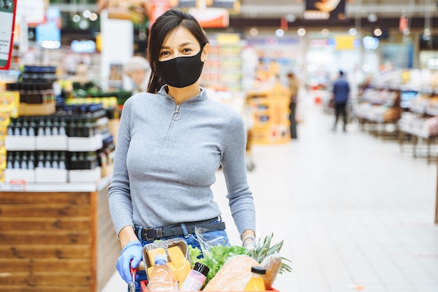 Foto retrato de uma jovem sorridente na máscara protetora e luvas segurando um carrinho de supermercado em um supermercado.