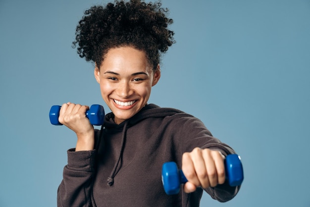 Retrato de uma jovem sorridente se exercitando com dois halteres posando isolados em fundo azul enquanto desfruta de treino e olha para a câmera