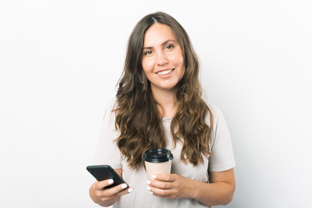 Retrato de uma jovem sorridente segurando um smartphone e uma xícara de café de papel