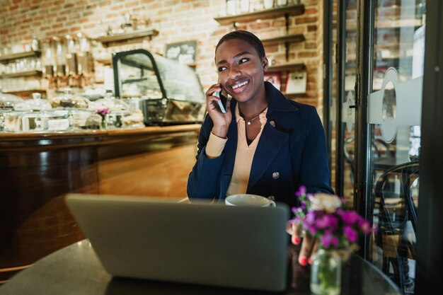 Foto retrato de uma jovem sorridente usando laptop e falando no telefone celular em uma cafeteria