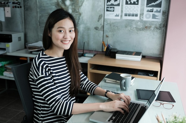 Retrato de uma jovem sorridente usando um laptop enquanto está sentada em uma mesa de escritório