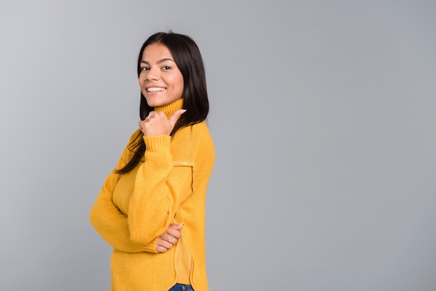 Retrato de uma jovem sorridente, vestida com um suéter, isolado na parede cinza, apontando para o lado oposto