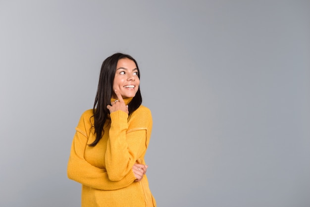 Retrato de uma jovem sorridente, vestida com um suéter, isolado sobre uma parede cinza, olhando para longe