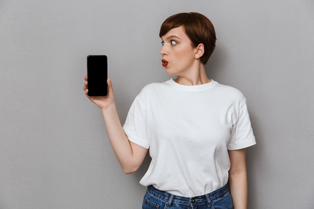 Retrato de uma jovem surpresa, sorrindo e segurando o celular isolado sobre uma parede cinza