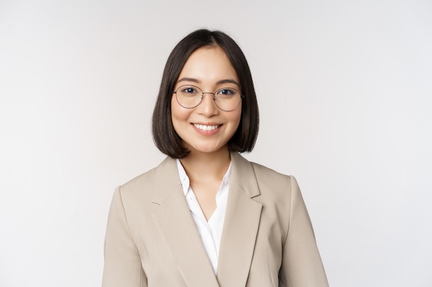 Retrato de uma jovem vendedora asiática em óculos vestindo terno bege sorrindo e parecendo confiante no fundo branco da câmera