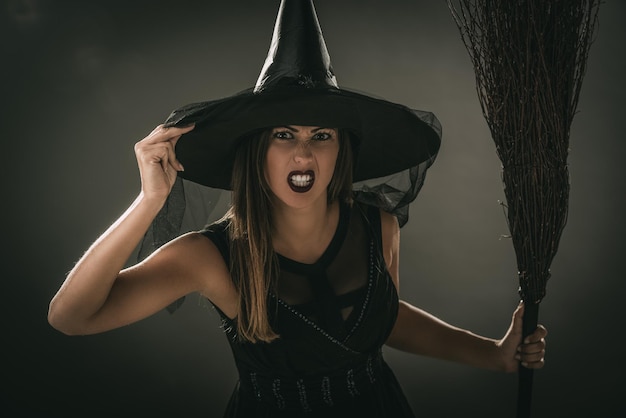 Retrato de uma jovem vestida como uma bruxa. Ela está com roupas escuras e segurando uma vassoura. Olhando para a câmera.