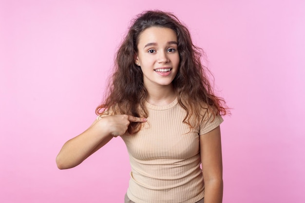 Retrato de uma linda adolescente com cabelo moreno encaracolado, vestindo roupas bege estilo casual, sorrindo para a câmera e apontando para si mesma, orgulhosa das conquistas. tiro de estúdio isolado no fundo rosa