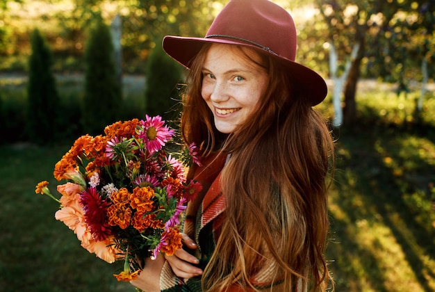 Retrato de uma linda adolescente ruiva com um chapéu vermelho segurando um buquê de flores ao pôr do sol