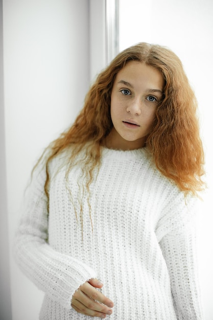 Retrato de uma linda adolescente ruiva em grande - suéter branco de malha em pé perto da janela no dia de inverno. mundo interior. Cabelo desgrenhado. Beleza natural. Diversas pessoas. Ternura e fragilidade.