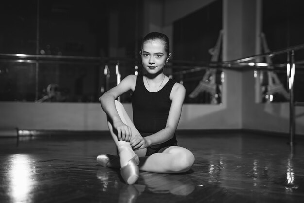 Retrato de uma linda bailarina sentada no chão e amarrando a ponta no estúdio de dança clássica. Menina sorridente, sonhando em se tornar dançarina de balé profissional. Retrato em preto e branco.