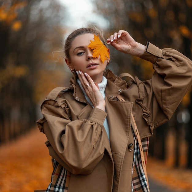 Foto retrato de uma linda e muito jovem mulher com roupas quentes elegantes e uma folha de bordo perto do rosto