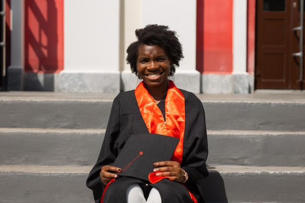 retrato de uma linda estudante africana em uniforme de formatura com um diploma nas mãos