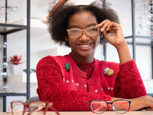 Retrato de uma linda garota africana adolescente com penteado afro usando um vestido vermelho está experimentando óculos em uma loja de óptica durante a temporada de Natal