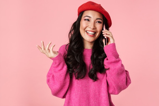 Retrato de uma linda garota asiática de boina sorrindo e falando em smartphone isolado em rosa