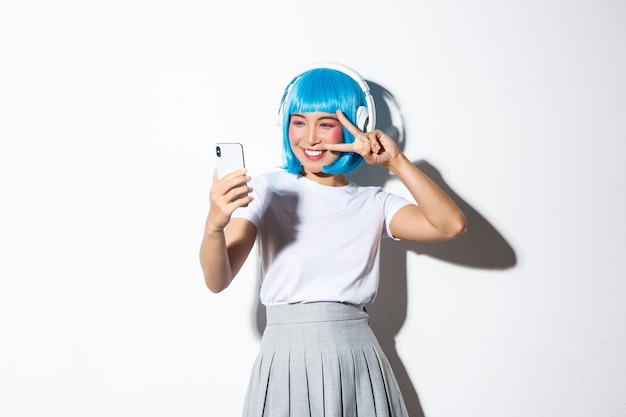 Retrato de uma linda garota asiática vestida de personagem de anime, posando para selfie com fones de ouvido e peruca curta azul