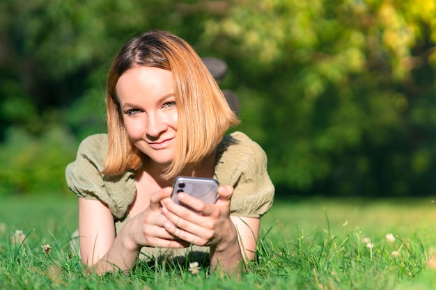 Retrato de uma linda garota atraente, jovem feliz alegre positiva mulher deitada no parque de verão na grama, sorrindo e olhando para a câmera com seu telefone celular, smartphone
