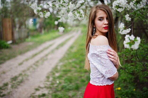 Retrato de uma linda garota com lábios vermelhos no jardim da flor da primavera usa vestido vermelho e blusa branca