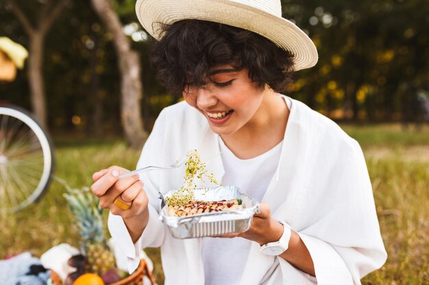 Retrato de uma linda garota de chapéu e camisa branca comendo comida deliciosa alegremente passando tempo no piquenique no parque