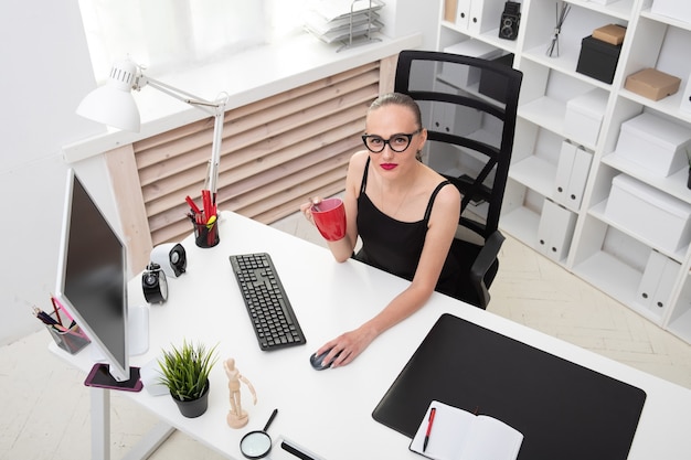 Retrato de uma linda garota em uma blusa preta que bebe café em uma caneca vermelha no local de trabalho