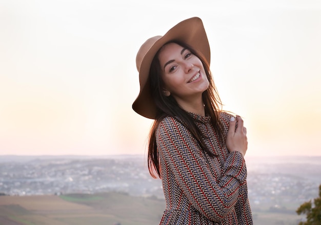 Retrato de uma linda garota feliz com um chapéu em um fundo de cidade ao pôr do sol. Vira-se para a câmera e dá um lindo sorriso brincalhão.