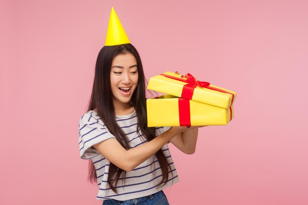 Retrato de uma linda garota feliz usando cone de festa espreitando dentro da caixa de presente com expressão espantada, agradavelmente surpreso e feliz em ver o melhor presente. tiro de estúdio interior isolado no fundo rosa