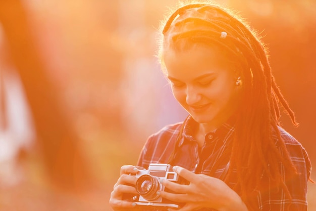 Retrato de uma linda garota hipster com dreads segurando a câmera e sorrindo no pôr do sol na vista ao ar livre da floresta