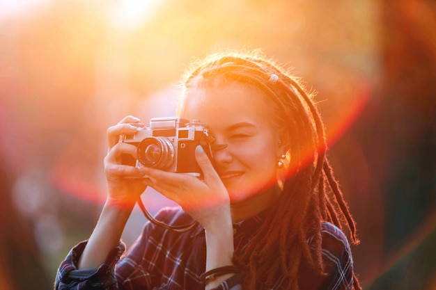 Retrato de uma linda garota hipster com dreads segurando a câmera e tirando fotos ao pôr do sol na vista ao ar livre da floresta